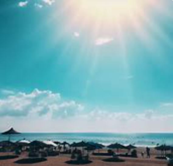 Imagen de una playa con sombrillas y personas bajo un cielo azul con el sol brillando.